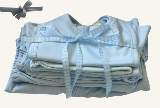 A stack of blue baby essentials including a sleep n play footie, pants, onesies, a hat, and a bib, all tied with a blue ribbon.